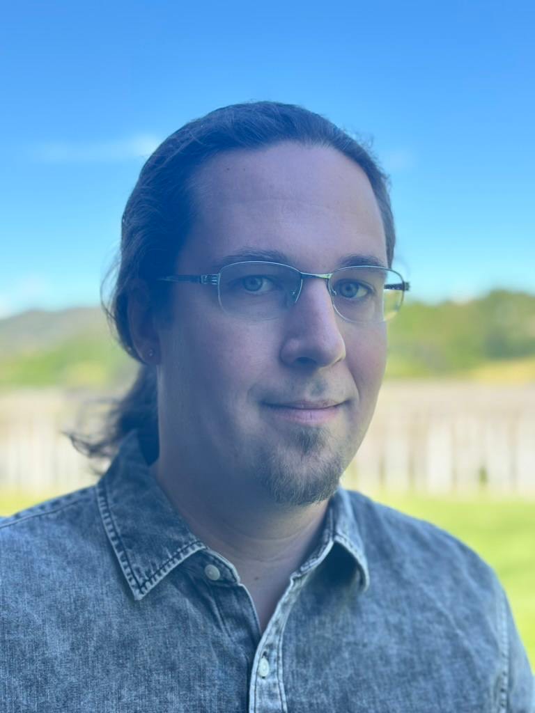 headshot of brandon hubler. 30's white male with glasses, goatee and ponytail, wearing black button up shirt. background is a blurry fenceline and mountain